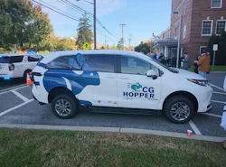 A white minivan with blue wave graphics and “CG Hopper by Via” branding, with a wheelchair accessibility symbol on its side, is parked in a lot beside a brick building, with a police vehicle with flashing lights and several people standing nearby.