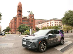 A gray and purple VIA Link Electric vehicle, unit 801R, with “Independent contractor driver” lettering, sits on a brick-paved street in front of a historic red sandstone courthouse with a tall tower and modern office buildings.