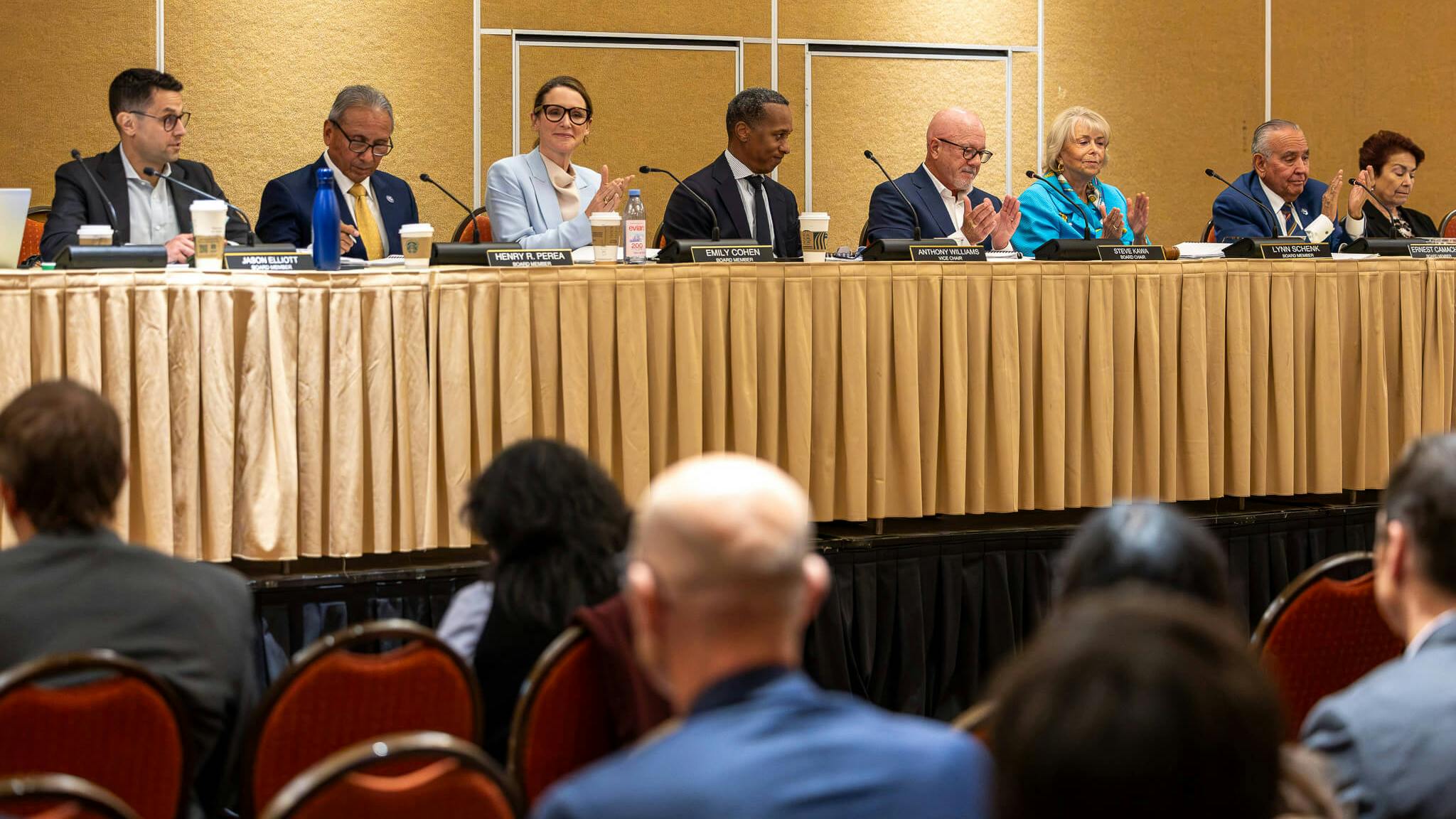 Board members Jason Elliott, Henry R. Perea, Emily Cohen, Anthony Williams, Steve Kawa, Lynn Schenk and Ernest Camacho sit at a long table during a meeting, with several applauding.